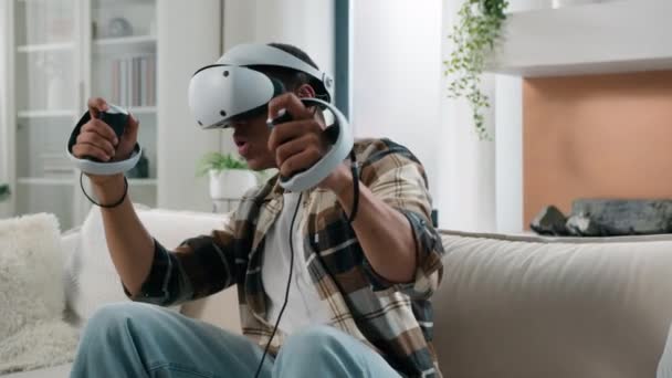 Hombre afroamericano en gafas de casco VR jugar videojuego realidad virtual experiencia metaverso jugar adicción a los juegos de ordenador divertirse en casa chico disfrutar del mundo cibernético 3d en línea con controladores - Metraje, vídeo