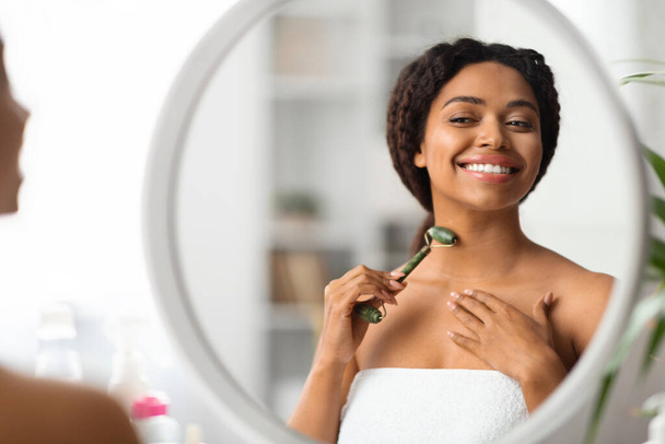 Linda jovem mulher negra massagem pescoço com Greenstone Jade Roller em casa, sorrindo Millennial afro-americano feminino olhando para o espelho e usando a ferramenta de beleza para cuidados com a pele, close-up - Foto, Imagem