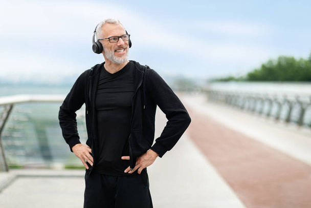 Healthy lifestyle for older adults. Cheerful athletic elderly man in black sportswear have workout outdoors, looking at copy space and smiling, using wireless headphones, listening to music - Foto, Imagen
