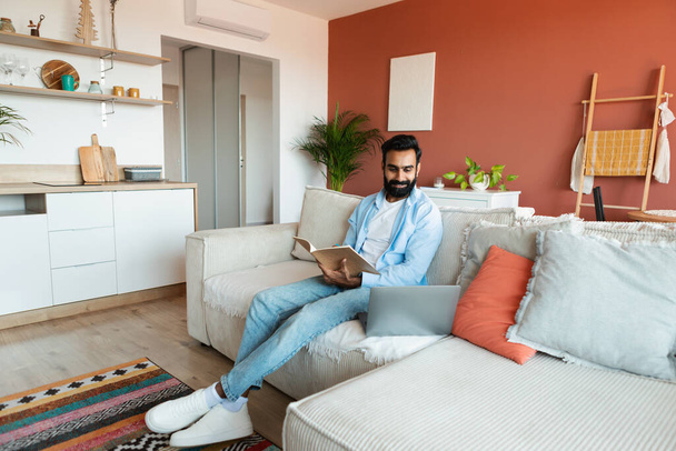 Happy intialainen mies käyttää kannettavaa tietokonetta ja lukeminen oppikirjan aikana online-oppitunnin opiskelu verkossa kotona, istuu sohvalla modernissa olohuoneessa. Kaveri työskentelee ja oppiminen etänä tietokoneen ja kirjan - Valokuva, kuva