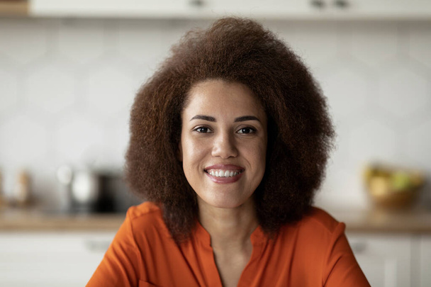 Nahaufnahme einer jungen glücklichen Afroamerikanerin mit buschiger Frisur, die im Hausinneren posiert, Porträt einer schönen schwarzen Millennial-Frau, die sich in der Küche entspannt, in die Kamera blickt und lächelt - Foto, Bild