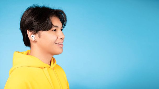 Koreaanse tiener jongen dragen oordopjes luisteren naar muziek, zijaanzicht portret over blauwe achtergrond. Aziatisch tiener ontspannen en genieten favoriete lied kijken naast op vrije ruimte, panorama - Foto, afbeelding