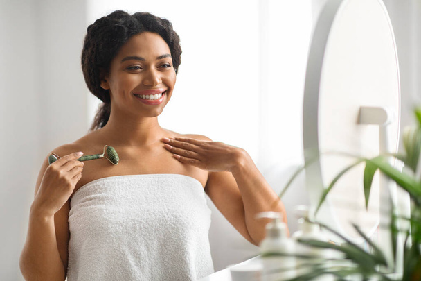 Tratamentos Skincare. Mulher afro-americana bonita usando Greenstone Jade Roller em casa, feliz preto feminino fazendo massagem de levantamento de corpo com ferramenta de beleza e olhando para o espelho, espaço livre - Foto, Imagem