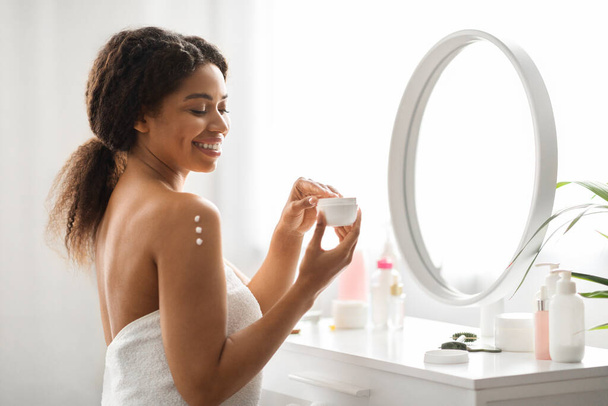 Θρεπτικό δέρμα. Ελκυστική μαύρη γυναίκα Εφαρμόζοντας ενυδατική κρέμα ή λοσιόν στον ώμο, χαμογελώντας όμορφη αφρο-αμερικανική κυρία τυλιγμένη σε πετσέτα μετά το μπάνιο κάνοντας θεραπείες ομορφιάς στο σπίτι - Φωτογραφία, εικόνα