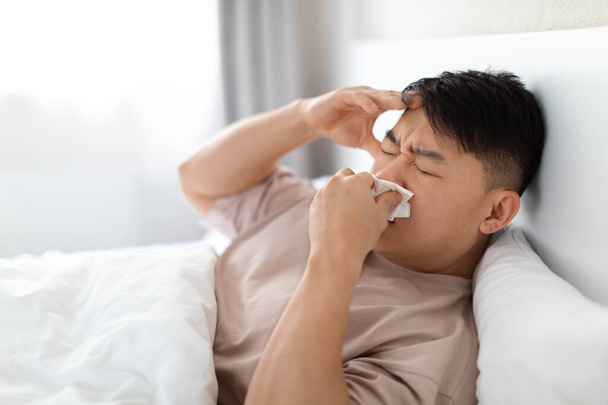 Malade homme asiatique d'âge moyen en pyjama couché au lit, touchant la tête, éternuant, souffrant de rhume, grippe, maux de tête, écoulement nasal, coronavirus, vue latérale, espace de copie - Photo, image