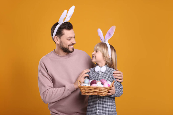 Father and son in bunny ears headbands with wicker basket of painted Easter eggs on orange background - Φωτογραφία, εικόνα