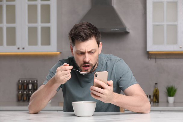 Man using smartphone while having breakfast at table in kitchen. Internet addiction - 写真・画像