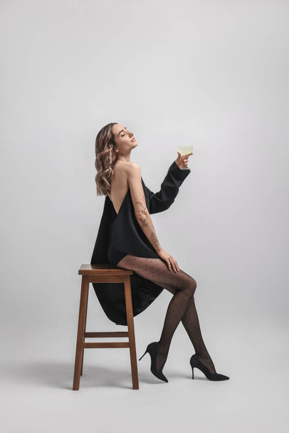 Όμορφη γυναίκα σε κομψό μαύρο ντύσιμο με ποτήρι ποτό ποζάροντας σε λευκό φόντο - Φωτογραφία, εικόνα