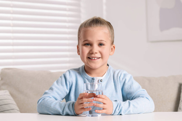 Happy little girl holding glass of fresh water at white table indoors - Foto, Bild
