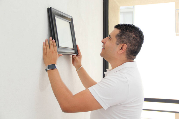 Σκούρα μαλλιά 40-year-old Latino άνθρωπος διακοσμεί το νέο του διαμέρισμα που αγόρασε με ένα μεσιτικό γραφείο, κρέμεται ένα πλαίσιο στον τοίχο - Φωτογραφία, εικόνα
