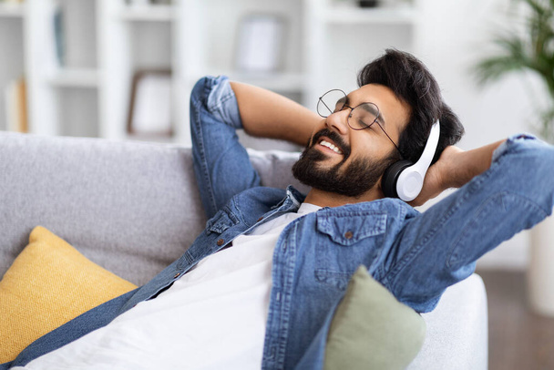 Guapo barbudo indio escuchando música en auriculares inalámbricos en casa, sonriendo joven oriental recostado en el sofá con los ojos cerrados y las manos detrás de la cabeza, disfrutando de la lista de reproducción favorita - Foto, Imagen
