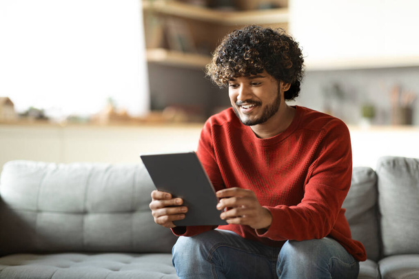 Handsome Indian Guy Spending Time With Digital Tablet At Home, Reading News, Browsing Internet, Watching Videos, Checking Social Networks While Relaxing On Couch In Living Room, Copy Space - Foto, Imagem