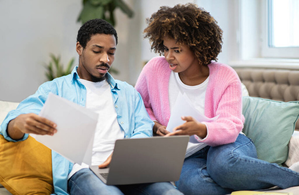 Mortgage problems concept. Overwhelmed young african american couple sitting together on couch, using laptop, holding papers documents, filling online form from home, have difficulties - Φωτογραφία, εικόνα