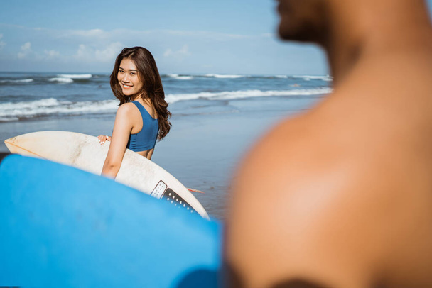 female surfer looks up with a smile to see a man carrying a surfboard on the beach - Photo, image