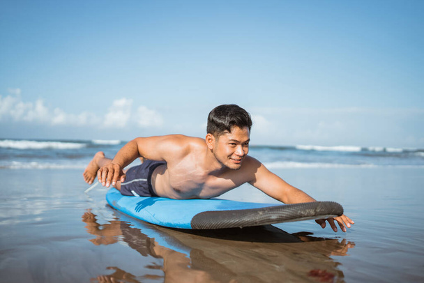 asiático macho surfista paletas en un surfboard en la arena antes de entrenamiento en la playa - Foto, imagen