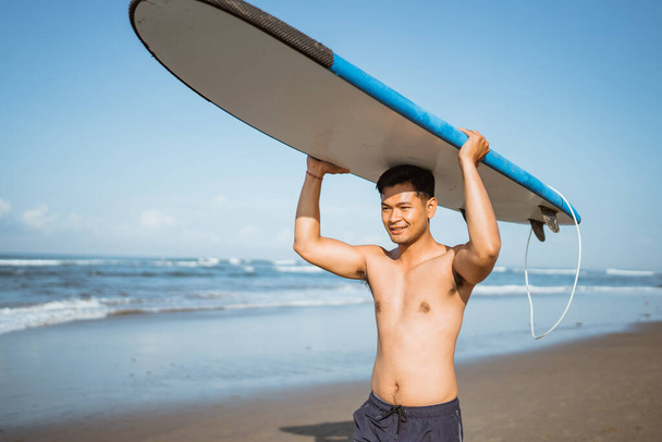 Smiling Asian man with a surfboard on his head walking on the beach enjoying a vacation - Foto, Imagem
