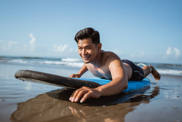 handsome young male surfer rowing on a surfboard while practicing on the sand at the beach - Photo, image