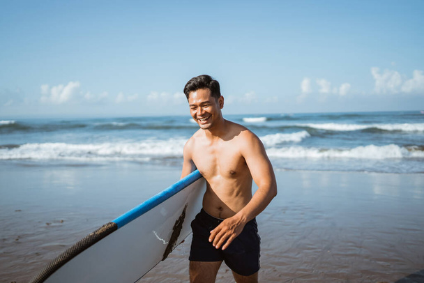 Asian male surfer happily carrying a surfboard after training during a beach walk - Photo, image
