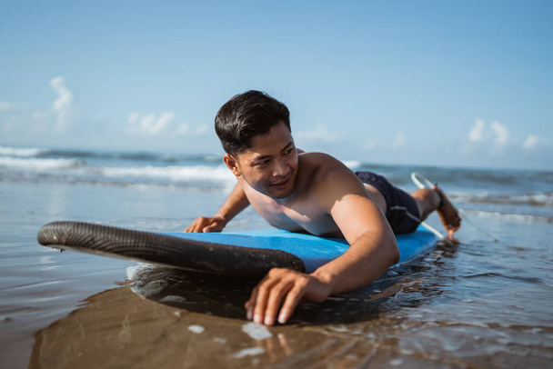 joven surfista remo en una tabla de surf mientras practica en la arena en la playa - Foto, Imagen