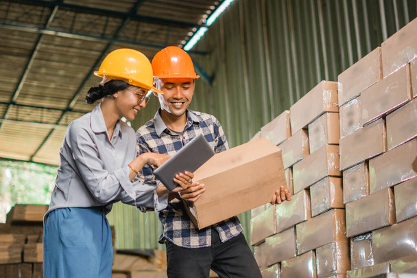 два заводські працівники носять безпечні шоломи під час використання планшета та картонних коробок на складі зберігання товарів - Фото, зображення