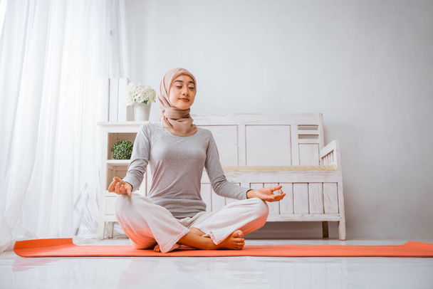 healthy muslim woman wearing hijab doing yoga pilates meditating in a room on orange mattress with eyes closed lotus pose in a room - 写真・画像