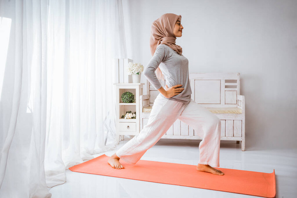 muzułmańskie hidżab kobieta rozgrzewka przed ćwiczeniami do domu poćwiczyć w pokoju z uśmiechem na pomarańczowym materacu - Zdjęcie, obraz