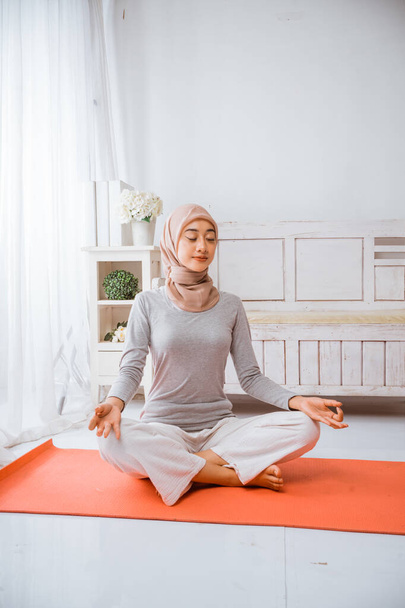 υγιή μουσουλμάνα γυναίκα φορώντας χιτζάμπ κάνει πιλάτες γιόγκα διαλογισμό σε ένα δωμάτιο σε πορτοκαλί στρώμα με μάτια κλειστά λωτού θέτουν σε ένα δωμάτιο - Φωτογραφία, εικόνα