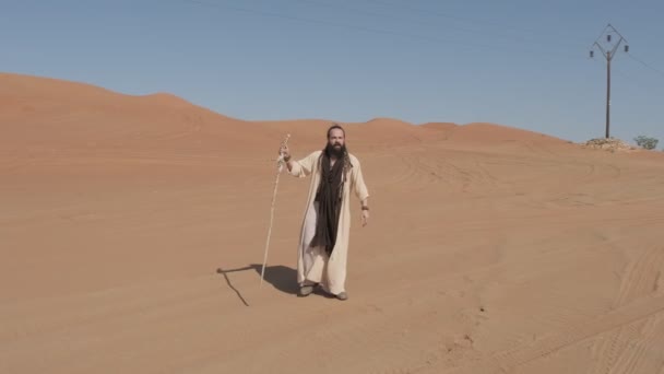 Um jovem com roupas de xamã branco com uma vara na mão caminha na areia do deserto contra o pano de fundo do pôr do sol. EAU em câmara lenta - Filmagem, Vídeo