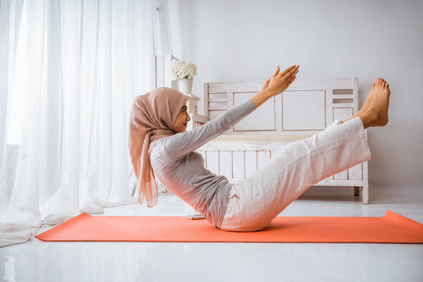 musulmana hijab mujer calentando antes de ejercicio haciendo yoga pilates pose en naranja colchón en un habitación - Foto, Imagen