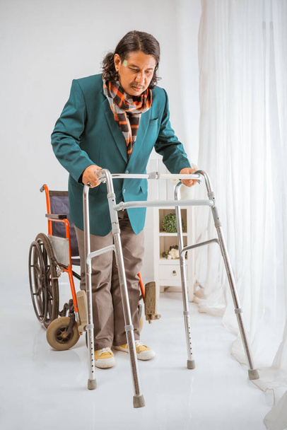 elderly man sitting on wheelchair trying to walk with walker frame in nursing home care - Foto, Imagen