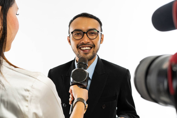 dorosły mężczyzna w niebieskiej koszuli i czarnym garniturze w okularach do powiek, przeprowadzony przez dziennikarza za pomocą czarnego mikrofonu i nagrany przez kamerę - Zdjęcie, obraz