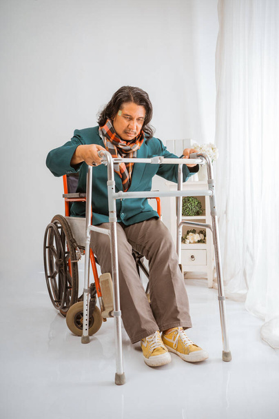 elderly man sitting on wheelchair trying to walk with walker frame in nursing home care - Fotó, kép