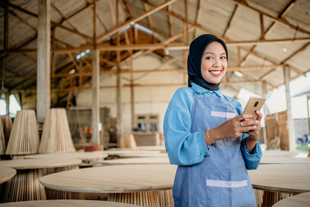smiling veiled businesswoman holding a cell phone while standing in a woodcraft shop - Foto, Imagen