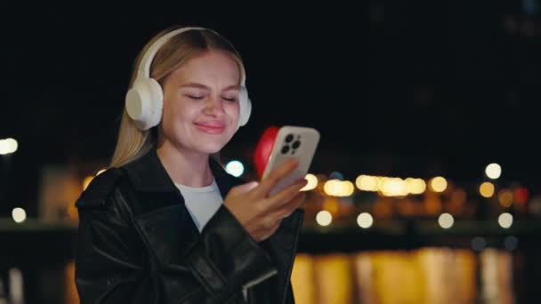 Jolie fille utilisant le téléphone portant des écouteurs blancs debout dehors la nuit. Young Lady À l'extérieur Écouter de la musique, utiliser un téléphone intelligent, envoyer des SMS, naviguer sur Internet. Concept Technologie et personnes - Séquence, vidéo