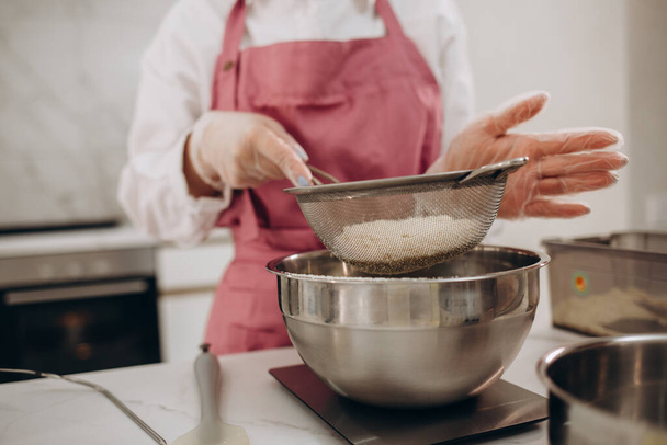 Une jeune femme blonde en chemisier blanc et pantalon beige prépare un gâteau dans la cuisine. Une femme verse de la farine dans un bol pour mélanger la crème. - Photo, image