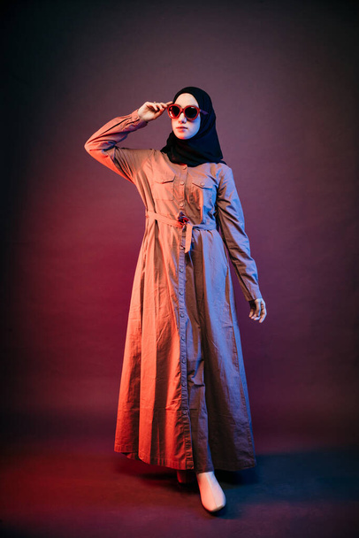 muslim woman wearing black hijab and orange long dress holding sunglasses looking sideways and crossing her legs - Foto, Imagem