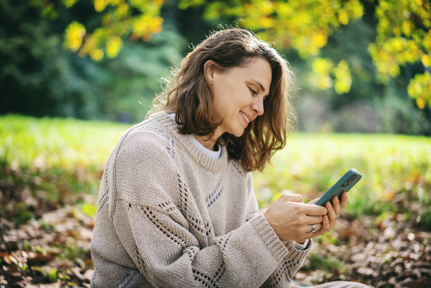 Charming curly young woman sitting in an autumn park with a mobile phone in her hands - Фото, изображение