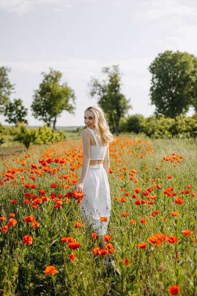 Blonde young woman standing in a field with wild red poppies, wearing a white dress, smiling, looking at camera. - Photo, image