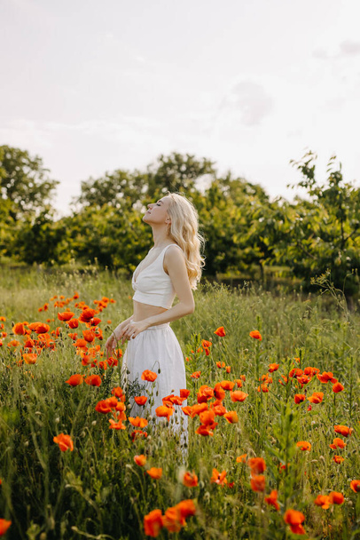 Blonde Frau steht auf einem Feld mit wilden roten Mohnblumen, trägt ein weißes Kleid und genießt die frische Luft. - Foto, Bild