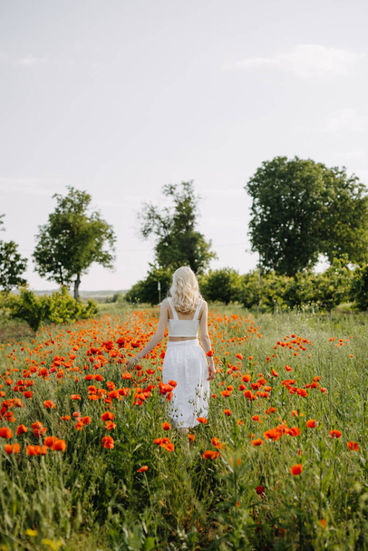 Blonde Frau geht durch ein Feld mit wilden roten Mohnblumen und trägt ein weißes Kleid. - Foto, Bild