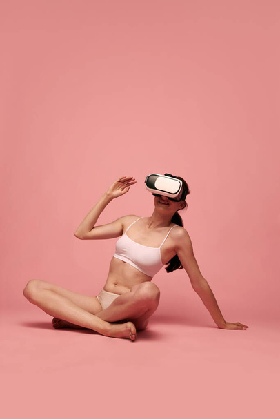 Young smiling woman with slim, fit body, wearing vr glasses, sitting on floor in underwear against pink studio background. Concept of female beauty, body care, fitness, sport, health, figure, ad - Foto, immagini