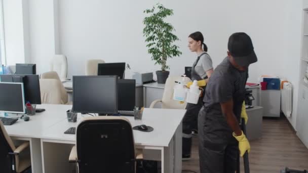 Scatto al rallentatore della giovane donna che porta detergenti alle scrivanie pulite nell'ufficio open space mentre il suo collega maschio usa l'aspirapolvere - Filmati, video