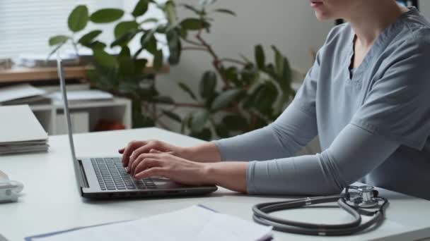 Fiatal, magabiztos női orvos vagy más, kék egyenruhás orvos közelsége laptopbillentyűzeten ülve, miközben a munkahelyen sztetoszkóppal a klinikai irodában ül - Felvétel, videó