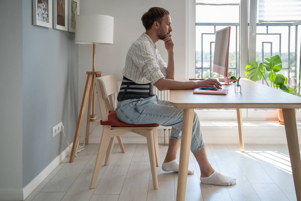 コンピュータを使用してテーブルに座ってルーバーブレースを着用する集中男性フリーランサーは,若い男性は拡声ディスクで遠隔操作コルセットベルトを使用しています. 自宅や背中の痛みからの仕事  - 写真・画像