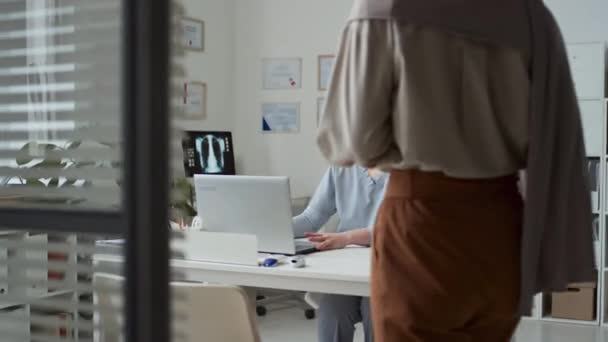 Mladý lékař v uniformě skládací notebook, zatímco muslimský žena pacientka v hidžábu vstupu do klinické kanceláře a sedí před ní před lékařskou konzultaci - Záběry, video