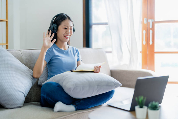 Asiatique jeune femme d'affaires est assis sur le canapé porte casque vidéoconférence avec ordinateur portable lever la main en disant salut équipe au bureau à domicile, travail de la maison - Photo, image