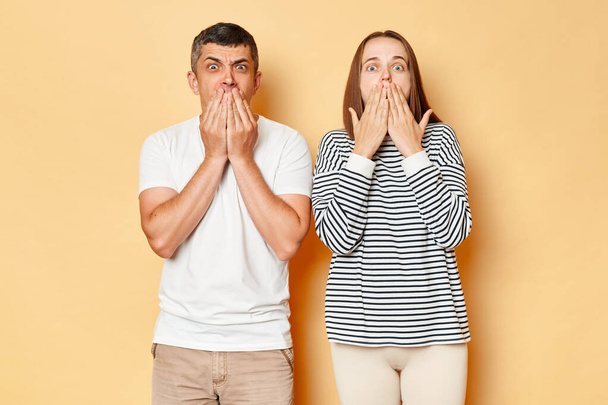 Shocked astonished young couple wife and husband wearing casual attires standing isolated over beige background looking at camera with big eyes covering mouths with hands. - Photo, image