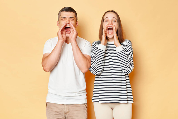 Excited amazed young couple wife and husband wearing casual attires standing isolated over beige background posing with hands near mouth screaming loud announcement. - Фото, изображение