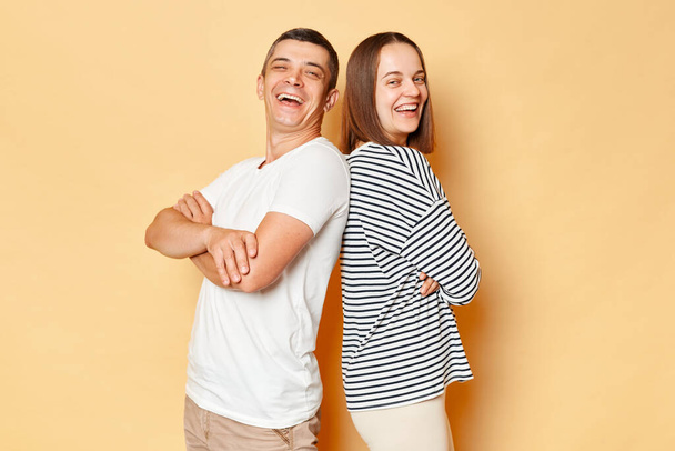 Happy cheerful young couple wife and husband wearing casual attires isolated over beige background standing back to back looking at cmaera and laughing having fun together. - Fotoğraf, Görsel