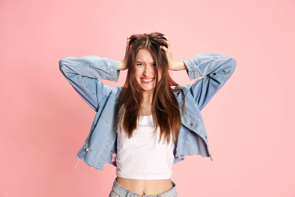 Portrait of young beautiful girl emotionally holding head with hands against pink studio background. Stress, chaos, irritation. Concept of human emotions, fashion, youth, lifestyle, female beauty, ad - Foto, immagini